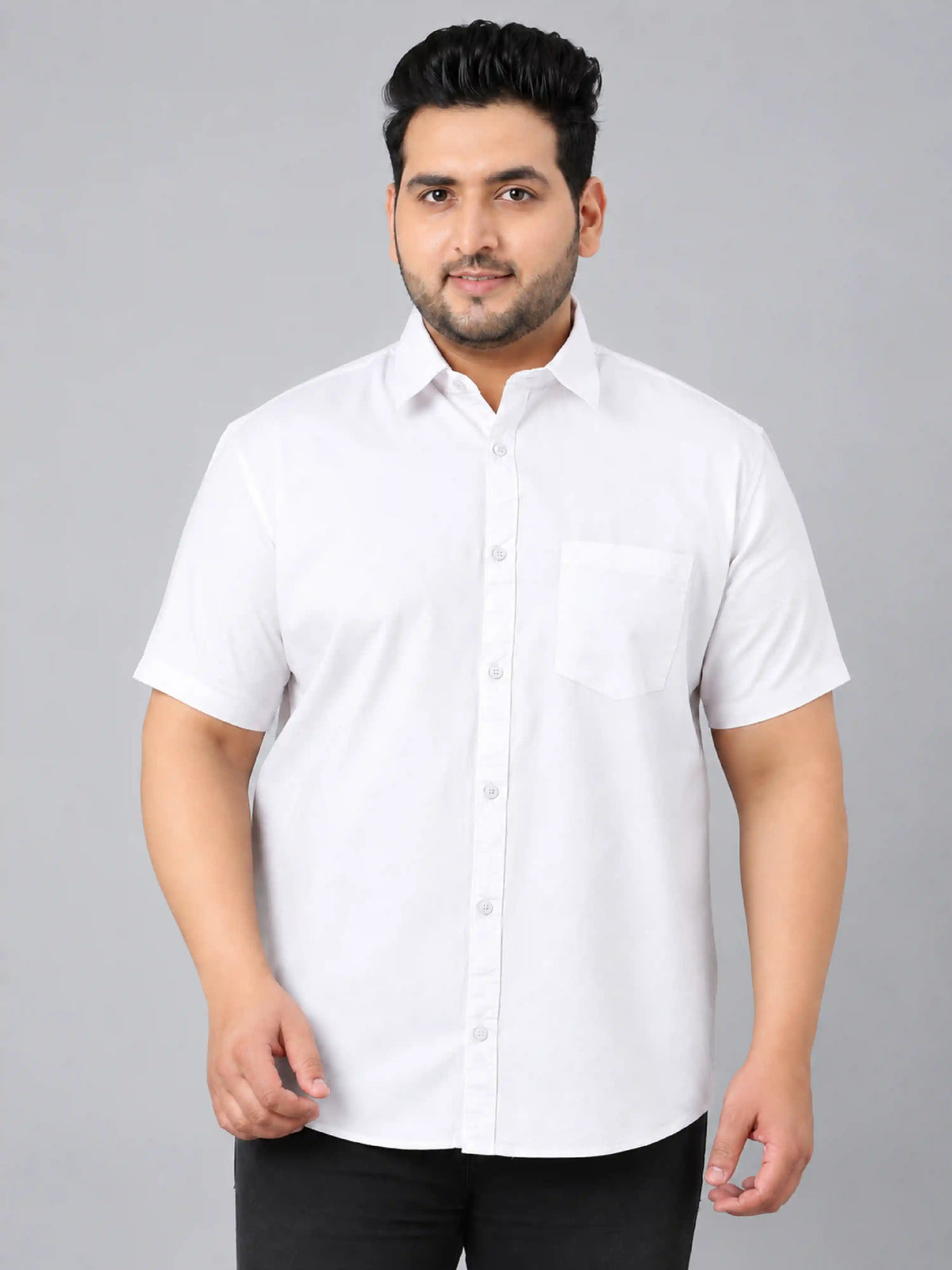 A man wearing a white short-sleeve button-up shirt with a chest pocket, paired with black trousers, standing against a solid gray studio background.