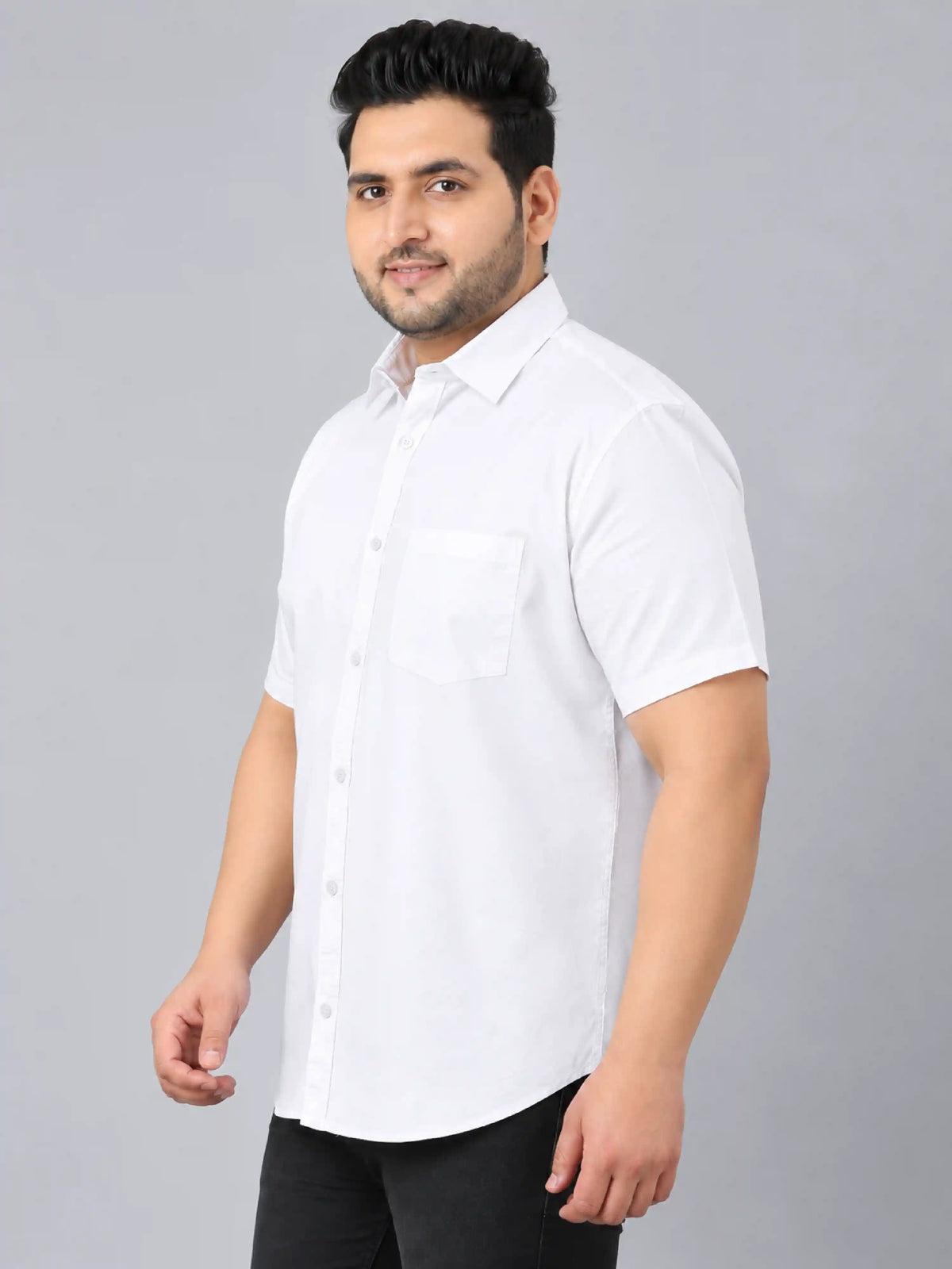 A man wearing a white short-sleeve button-up shirt with a chest pocket, paired with black trousers, standing against a solid gray studio background.
