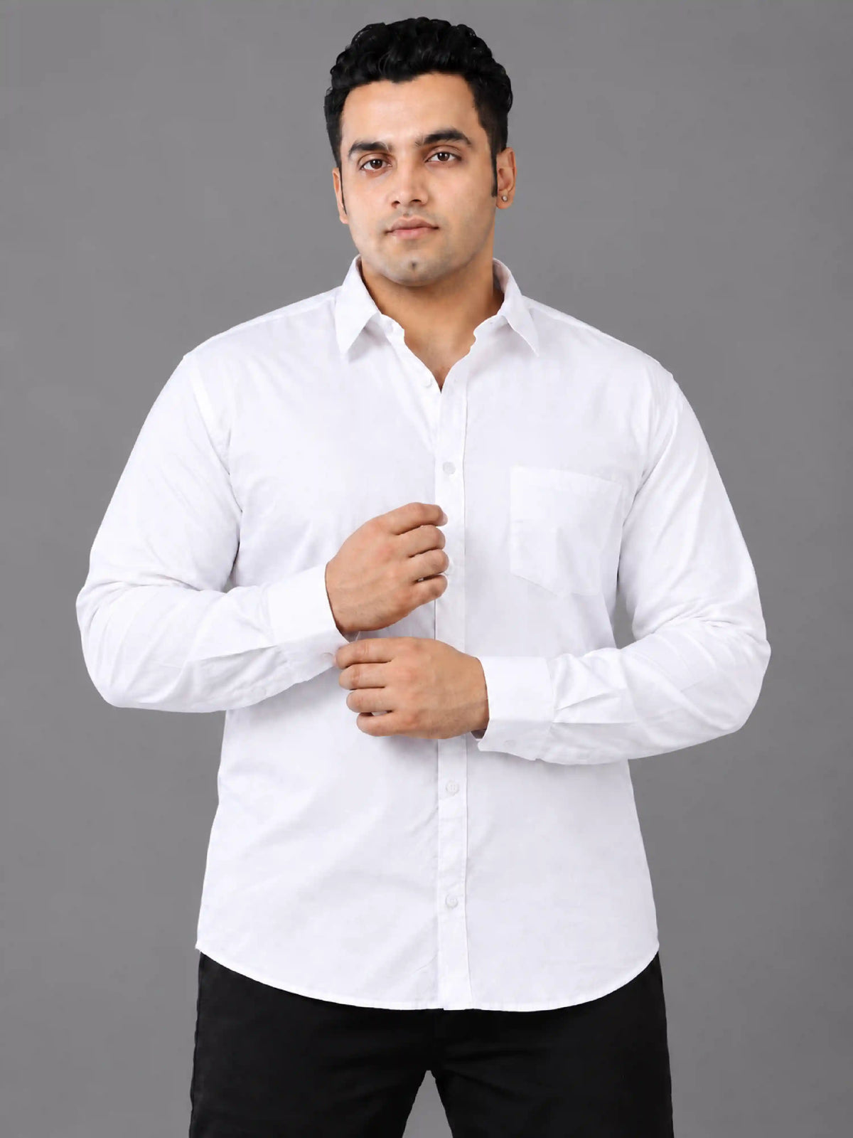 A man wearing a white long-sleeve button-down shirt with a chest pocket, paired with black trousers, standing against a solid gray studio background.